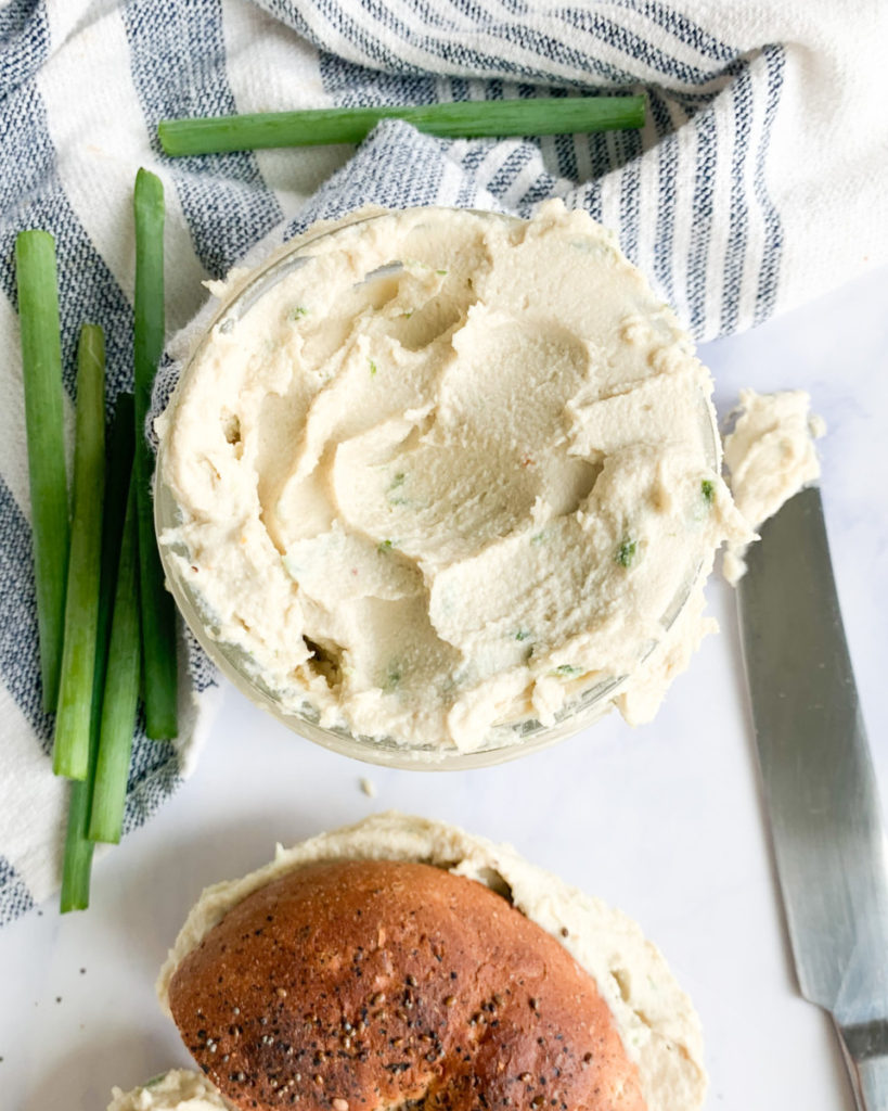 a vertical photo of a glass jar filled with vegan cashew cream cheese and half of a bagel on a white marble counter top 