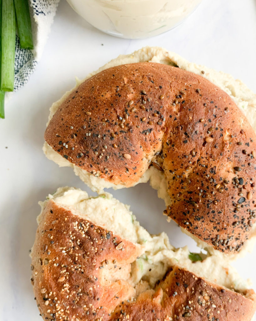 a vertical photo of a bagel with cream cheese that is cut in half on a white marble counter top 