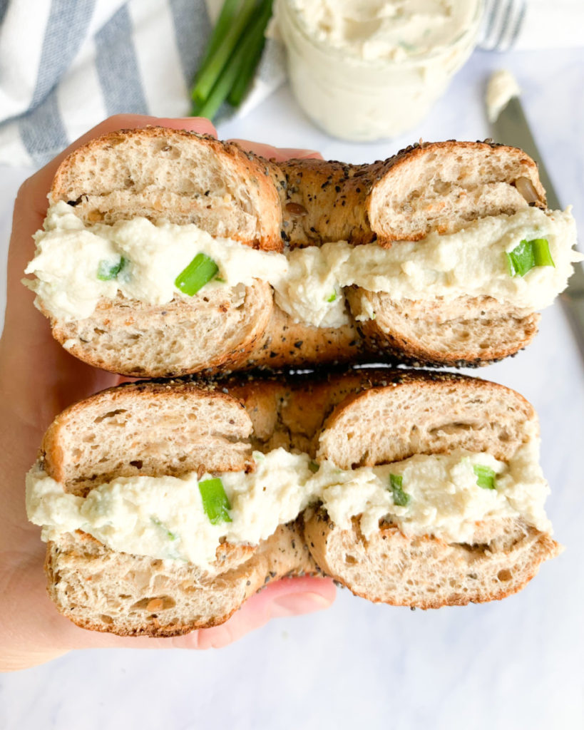 a vertical photo of a hand holding a bagel with vegan cashew cream cheese  on a white marble counter top 