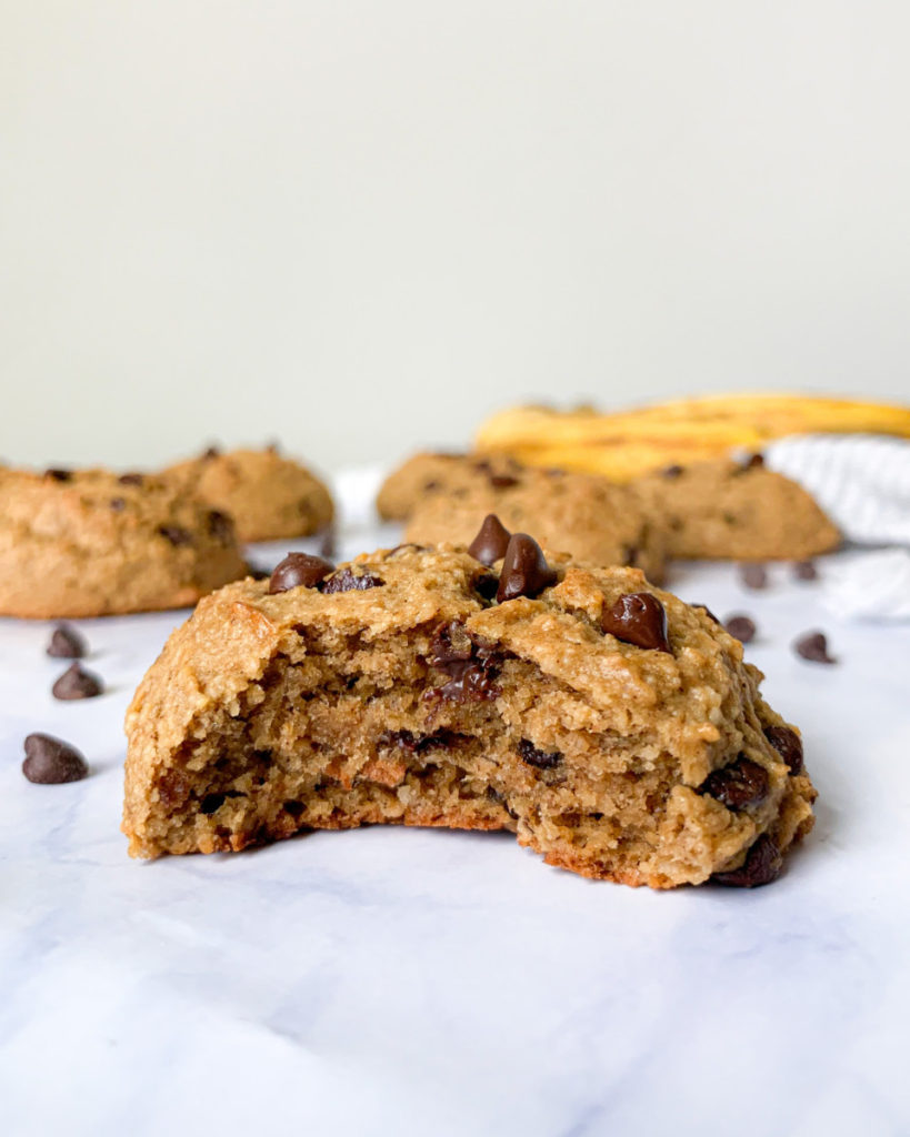 a vegan banana chocolate chip cookies on a white marble countertop with bananas and cookies in the background 