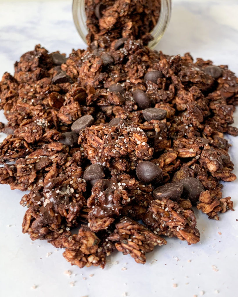 a vertical image of vegan salted chocolate probiotic granola spilling out of a mason jar onto a white counter top 