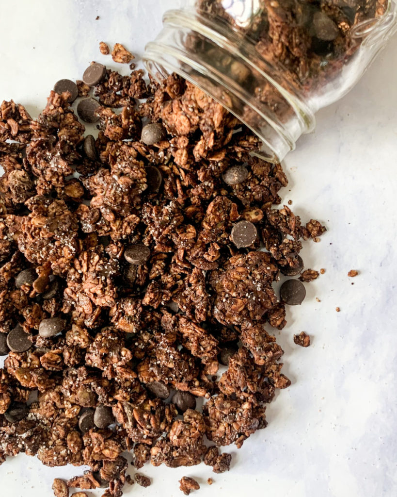 a vertical image of vegan salted chocolate probiotic granola spilling out of a mason jar onto a white counter top 