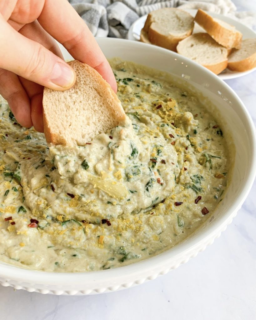 a piece of bread being dipped into a bowl of vegan spinach and artichoke dip with a plate of bread in the background