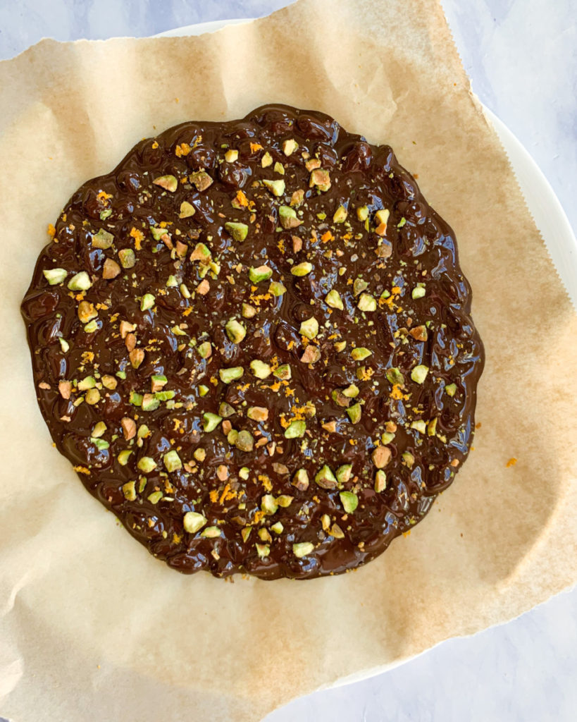 process photo of vegan dark chocolate pistachio and orange bark on parchment paper on a white plate on a white counter top 