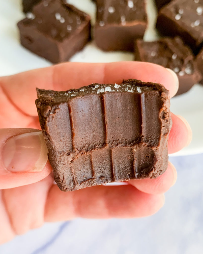 a vertical image of a hand holding a piece of oil free vegan dark chocolate peanut butter fudge with more fudge on a white plate in the background on a white counter top 