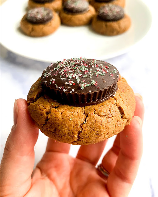 a vertical image of a hand holding a  flourless peanut butter cup blossom with cookies on a white plate in the background on a white counter top