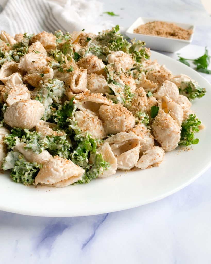 a vertical image of vegan creamy walnut pasta on a white plate with homemade vegan Italian breadcrumbs on a white counter top 