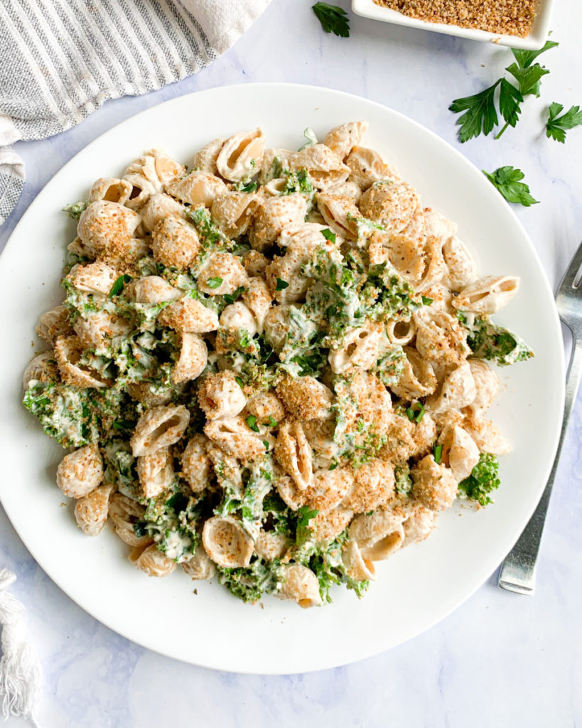 a vertical image of easy vegan creamy walnut pasta with homemade italian breadcrumbs on a white plate with a white and blue napkin and breadcrumbs in the background 