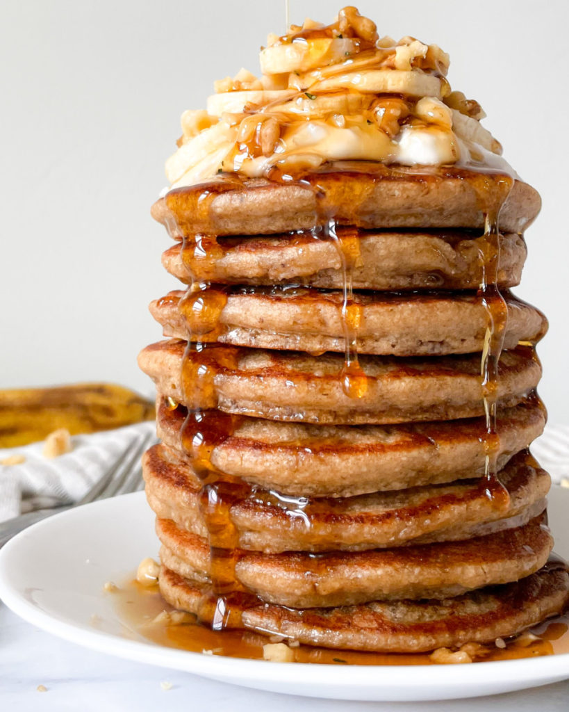 fluffy vegan banana pancakes stacked on a white plate with bananas walnuts and whipped cream on top with a napkin fork and banana in the background