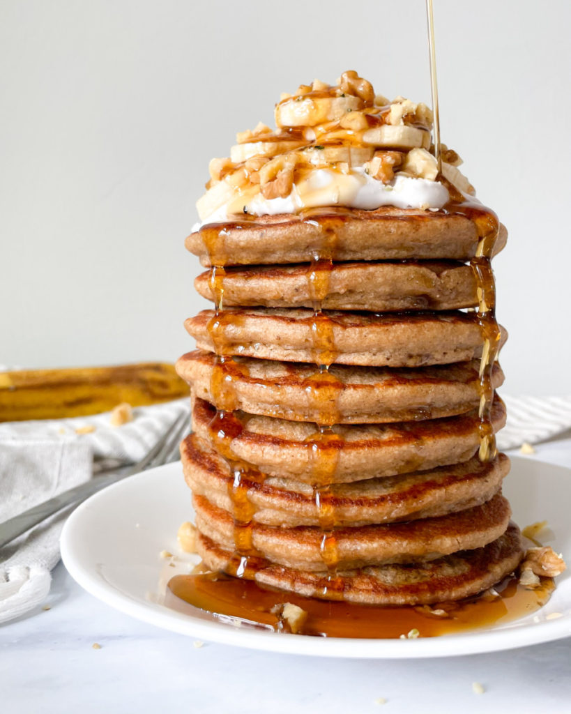 vegan blender banana oat pancakes stacked on a white plate with bananas walnuts and whipped cream on top with a napkin fork and banana in the background