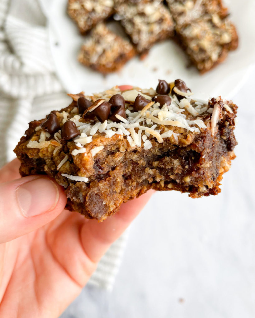 a vertical image of a hand holding vegan date sweetened oatmeal breakfast bars with more bars in the background on a white plate 
