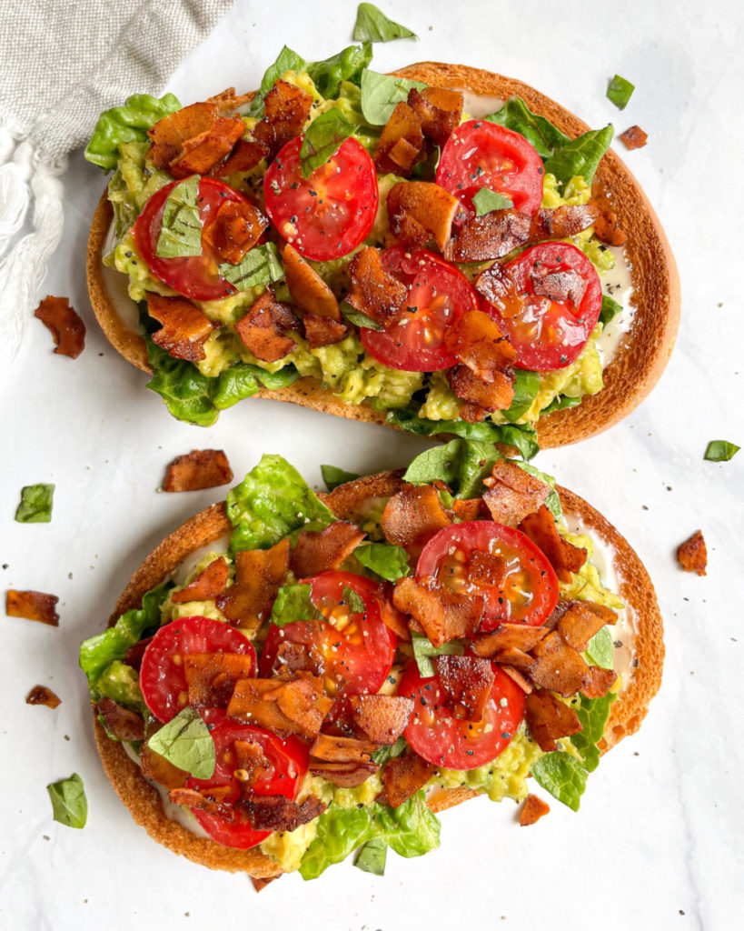 vegan coconut bacon on toast with avocado, vegan mayonnaise, romaine lettuce, tomatoes and basil on a white counter top  