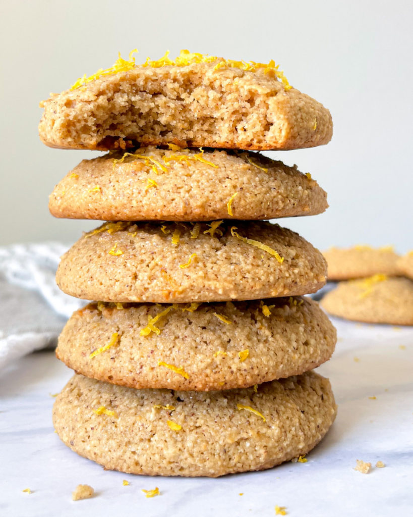 5 vegan lemon almond cookies stacked on a white counter top topped with lemon zest with more cookies in the background 