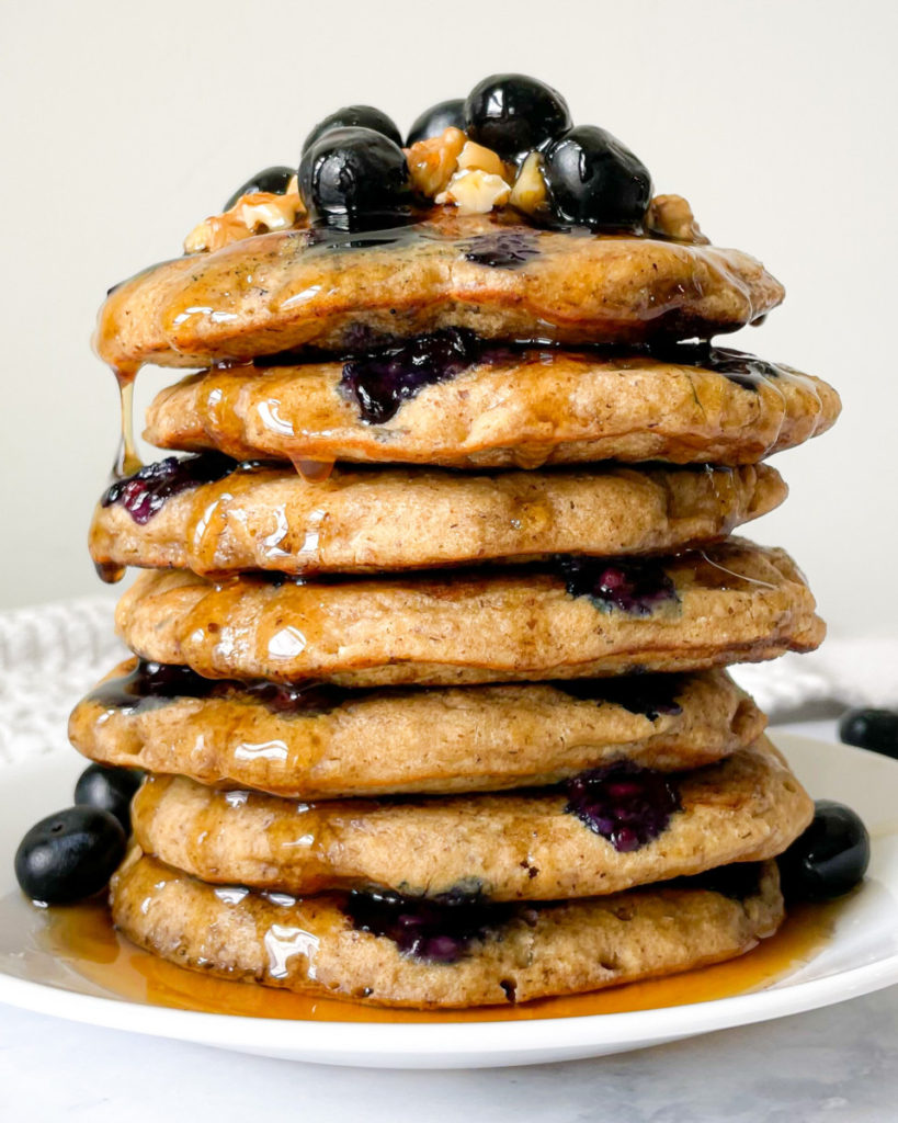 a close up photo of a stack of vegan blender blueberry pancakes with syrup, blueberries and walnuts on top 