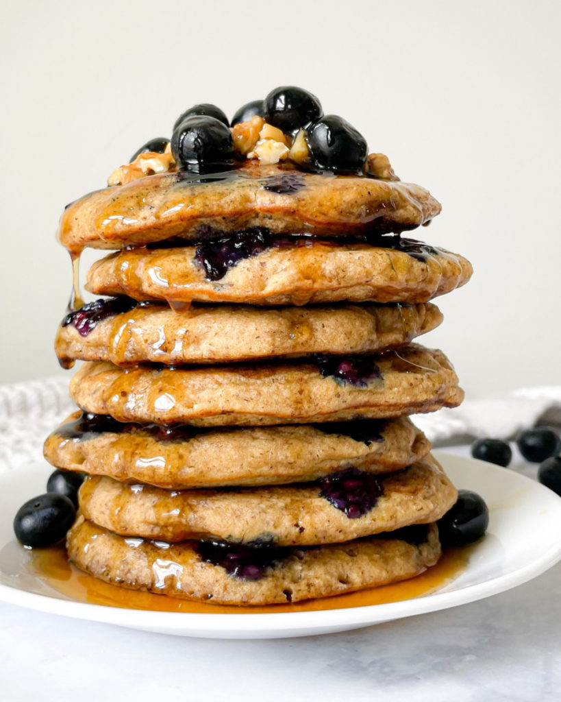 a vertical photo of a stack of vegan blender blueberry pancakes with blueberries and walnuts on top, maple syrup on a white plate on a white counter top 