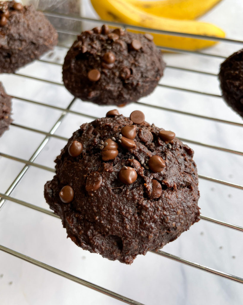 a close up photo of a vegan chocolate chia banana cookie on a cooling rack  on a white counter top with more cookies in the background 