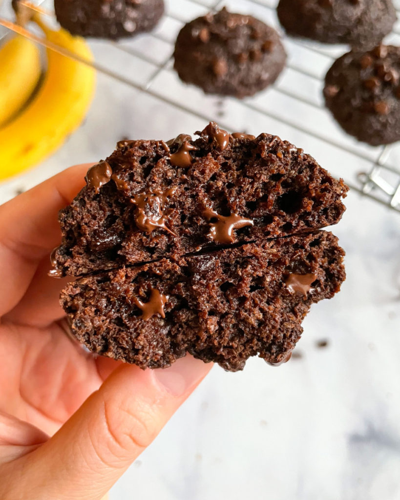 a hand holding a vegan chocolate chia banana cookie over a white counter top with bananas and more cookies on a cooling rack in the background