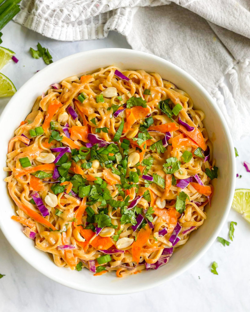 a big white bowl with vegan oil-free thai peanut noodles topped with carrots, purple cabbage, peanuts, green onions and cilantro in a white counter top 