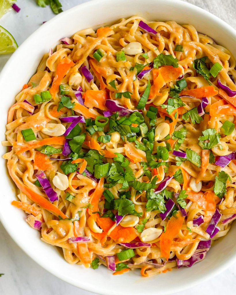 a close up photo of vegan oil-free thai peanut noodles with a vegan oil-free peanut sauce in a white bowl topped with veggies and cilantro on a white counter top 