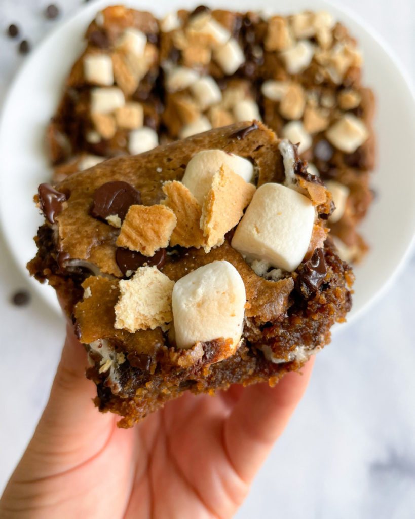 a hand holding a vegan peanut butter s'mores bar with a plate of s'mores bars in the background