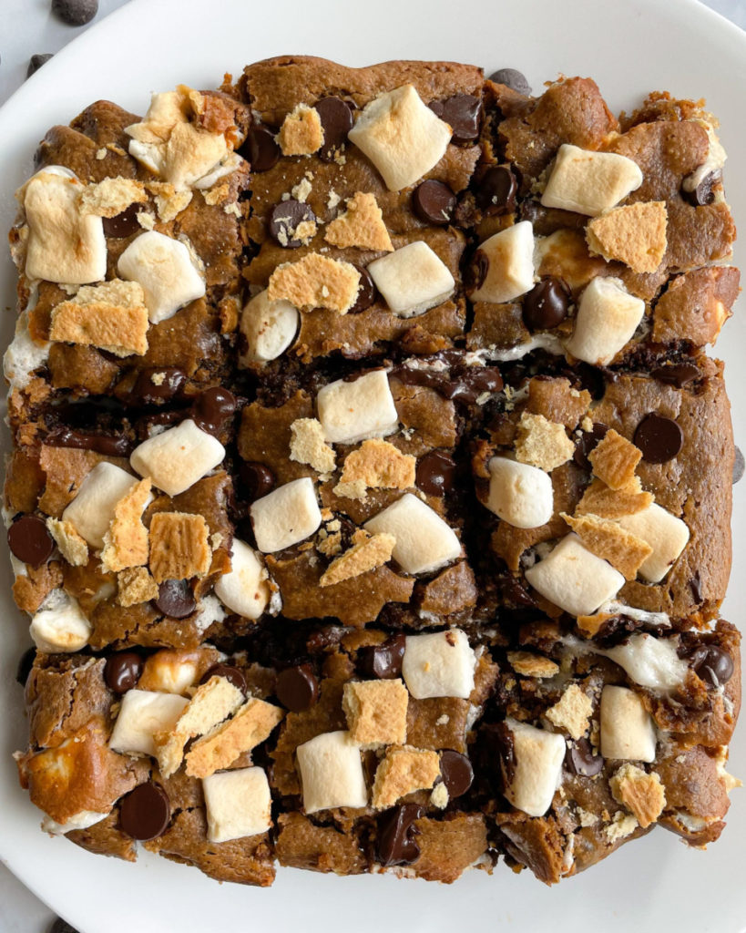 eight vegan peanut butter s'mores bars  on a white plate topped with marshmallows, dark chocolate chips and graham crackers 