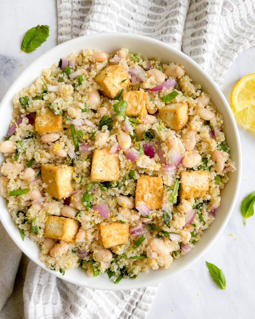 a big bowl of vegan white bean and quinoa salad with oil-free tofu on a white countertop with a gray and white napkin and basil and lemon on the counter 