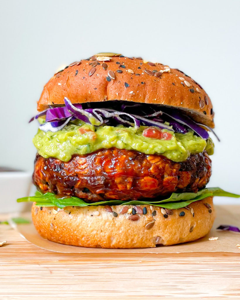 a close up photo of the best ever vegan BBQ veggie burger on bun with spinach, guacamole and purple cabbage on a wooden serving platter with BBQ sauce in the background 