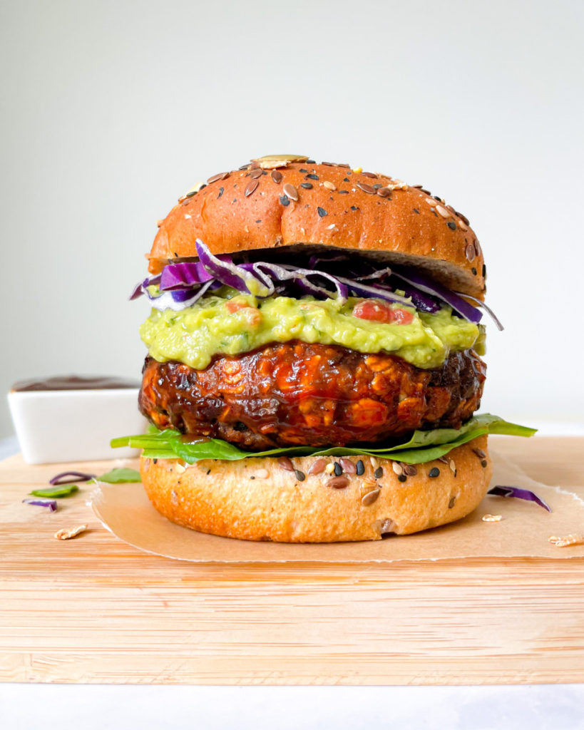 a photo of the best ever vegan BBQ veggie burger on bun with spinach, guacamole and purple cabbage on a wooden serving platter with BBQ sauce in the background 