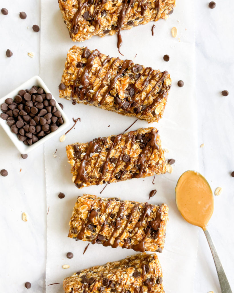 vegan date sweetened peanut butter granola bars with a chocolate drizzle on top on a white counter top next to a small bowl of mini vegan chocolate chips and a spoon with peanut butter 