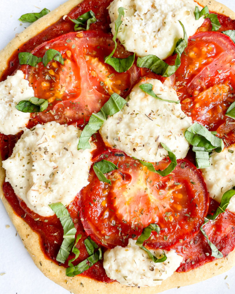 a close up photo of vegan oil-free chickpea and almond flour pizza crust with almond ricotta, tomatoes and basil